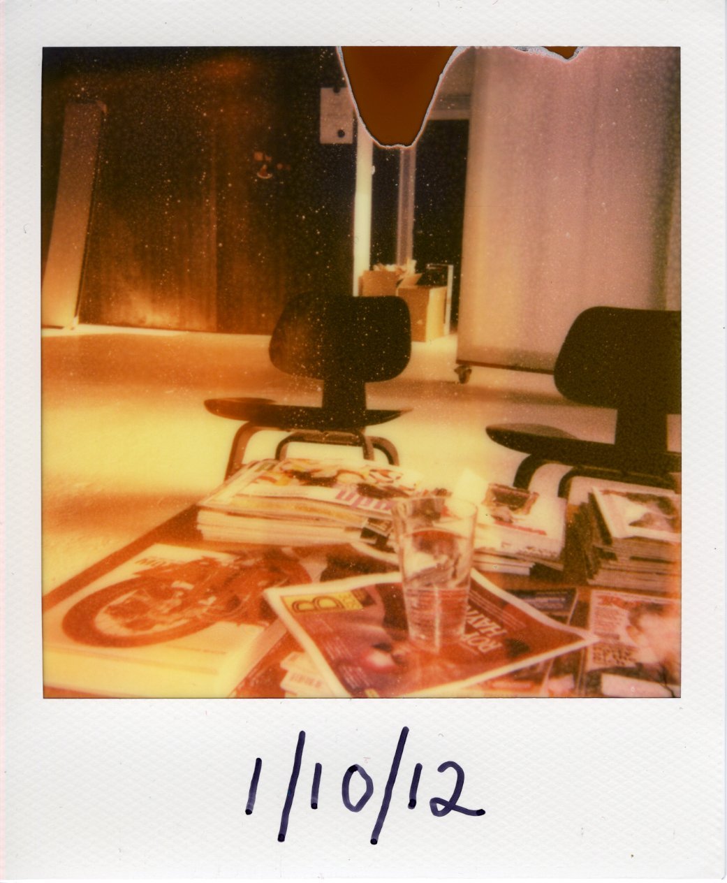 A sepia colored Polaroid photograph of an office lobby, there are chairs spread out and piles of newspapers and magazines with a glass of water placed on top of them.