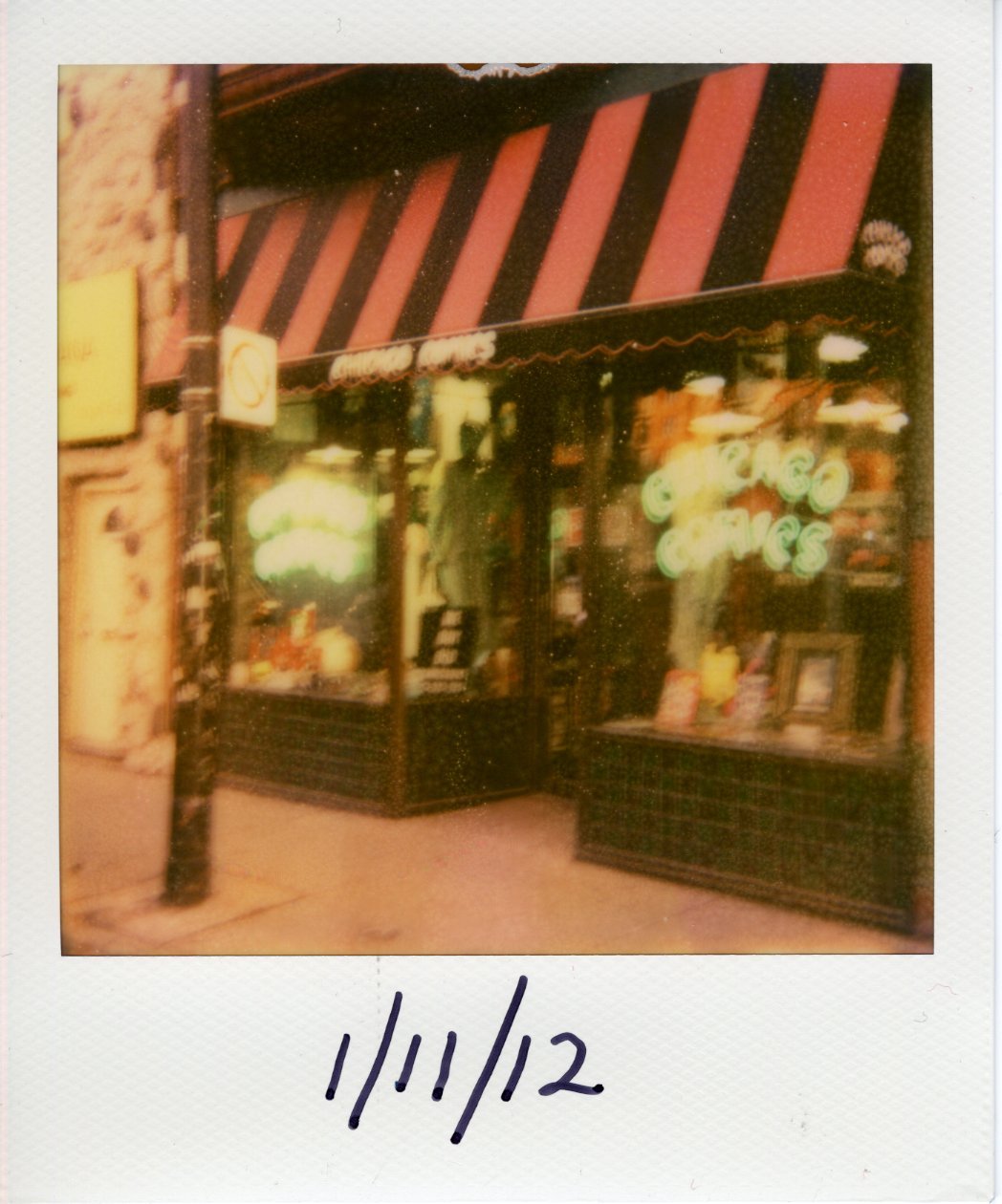 The busy window front of Chicago Comics complete with neon glowing logo signs and a red and black striped awning. We can see a utility pole directly in front of the door and many unidentifiable products laid out in the window.