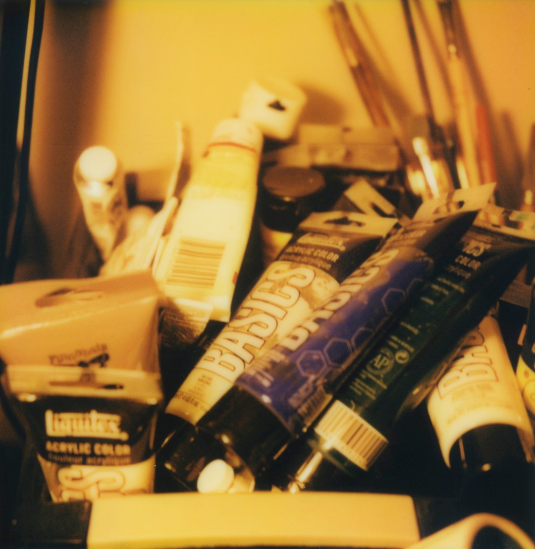 A large pile of disorganized acrylic paint tubes, on the right we can see a collection of brushes popping up.