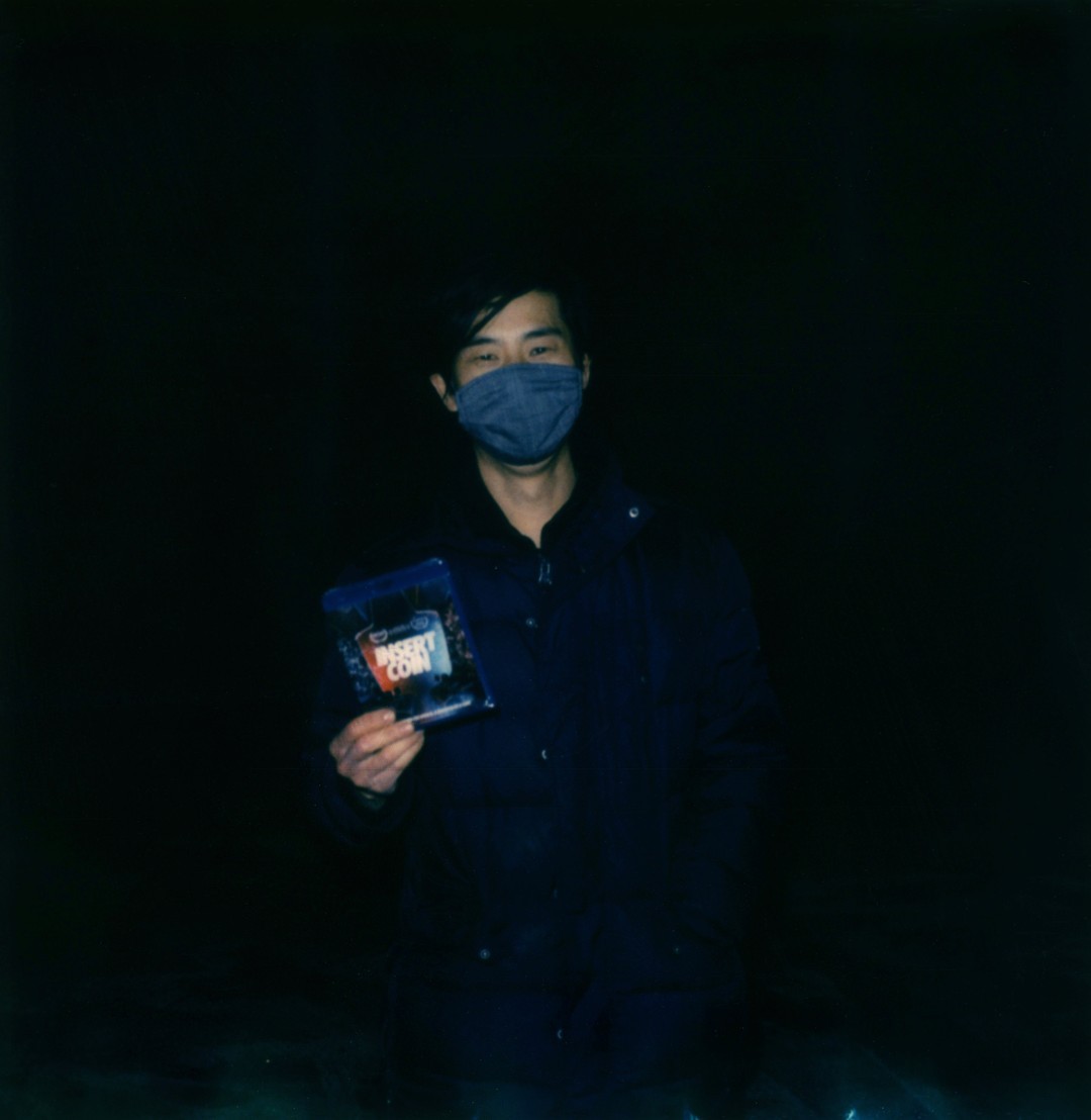 A man wearing a mask holding a Blu Ray disc of the film 'Insert Coin'