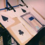 An easel with several pieces of white fabric laid out on them. Two prints show a black spade on them. One shows a black club.