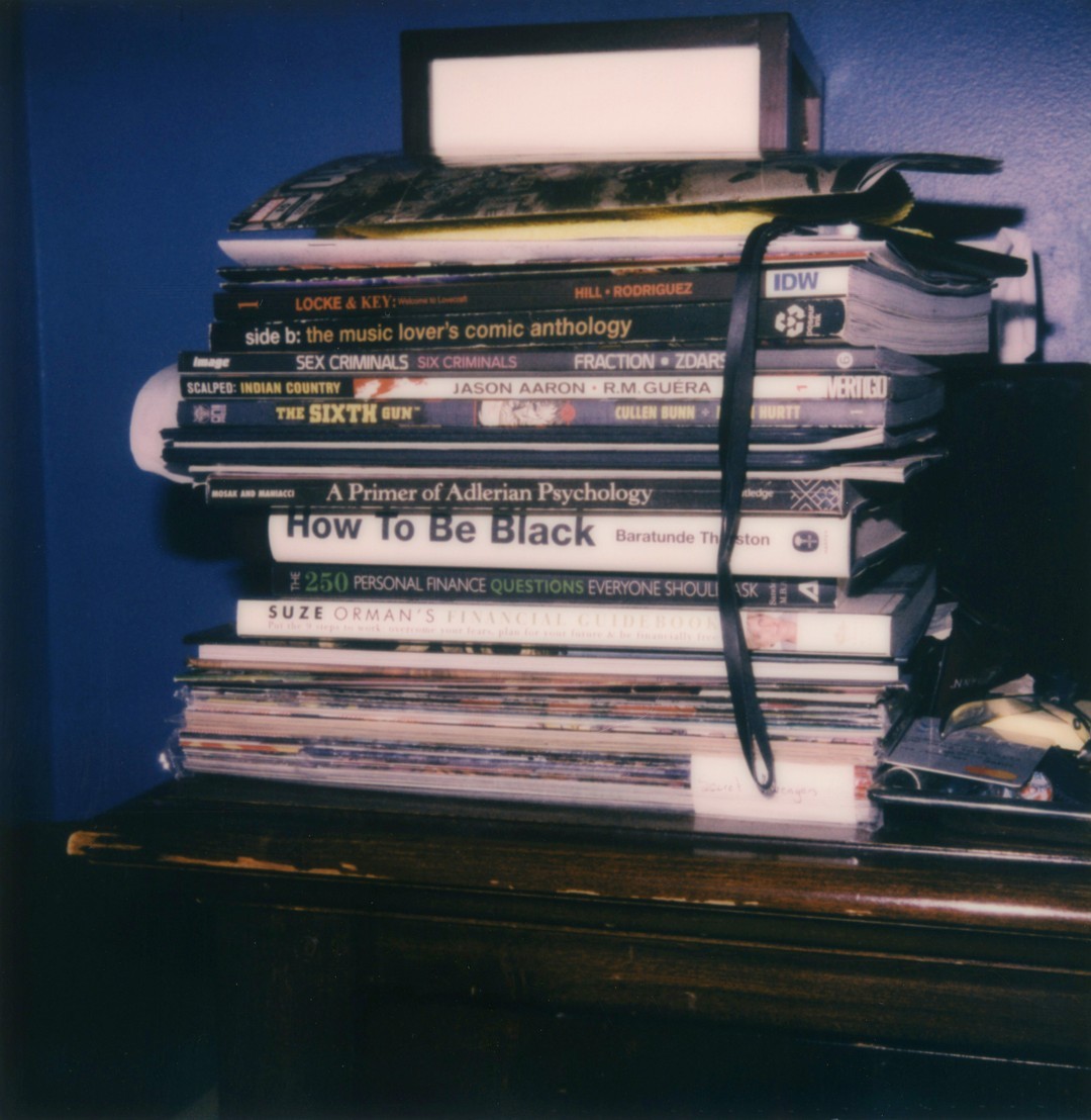 A big pile of books and comics on a nightstand.