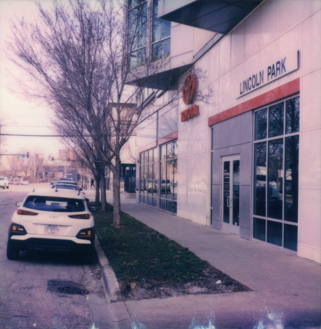 The entrance to a Toyota dealership. A car is parked in front and we can see a tree