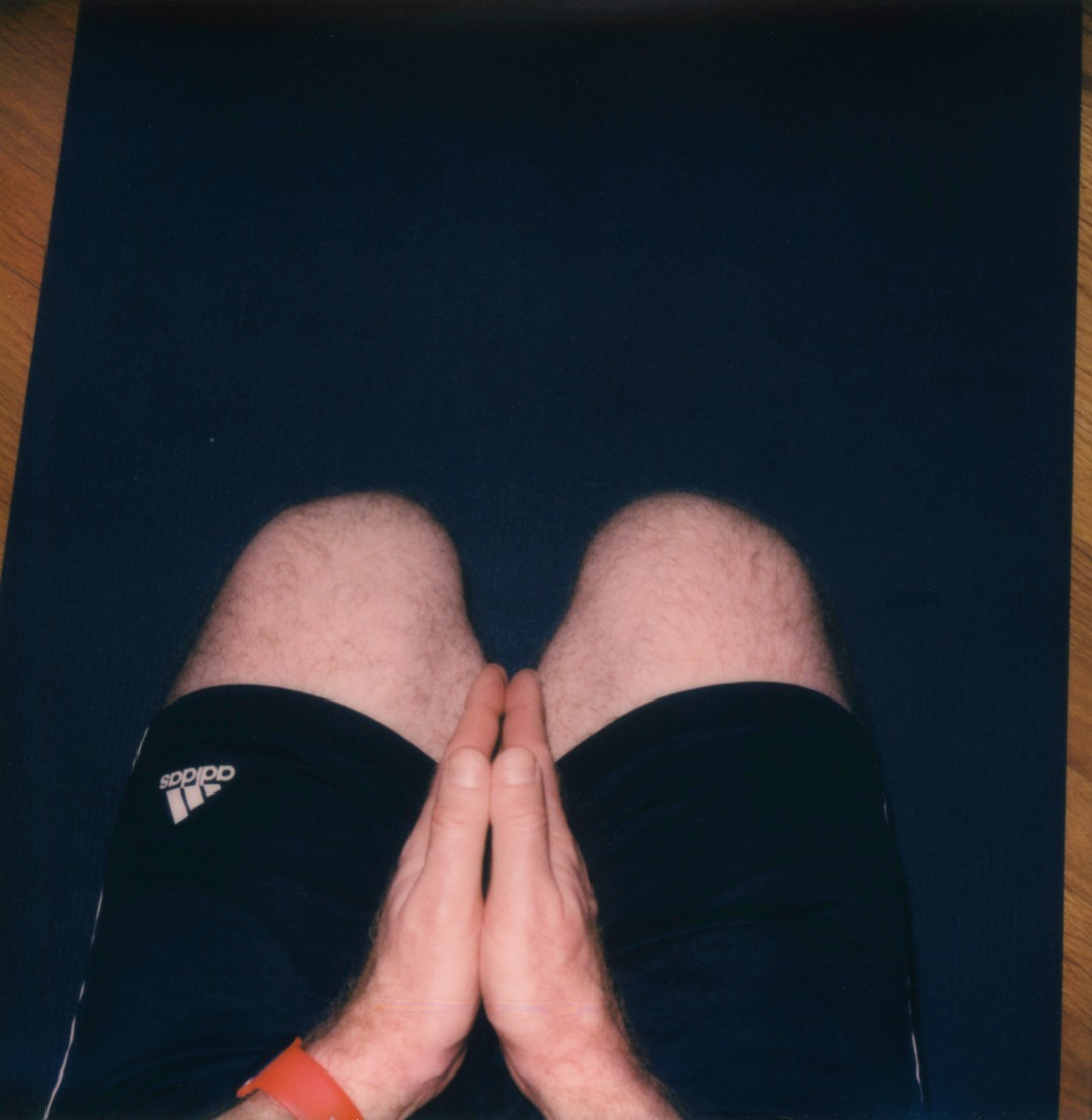 A top down view of a mans knees and hands on a yoga mat. He is wearing black Adidas shorts and his hands are in prayer position. He is wearing a red watchband.